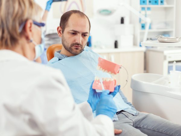 Stomatologist explaining proper dental hygiene using teeth skeleton during stomatology appointment. Dentist preparing sick man patient for dentistry surgery sitting on chair in examination room